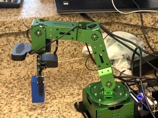 The green 6-DOF arm on a benchtop, gripper closed on a blue block.